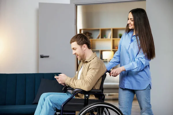 A cute young man in a wheelchair is holding a mobile phone while his girlfriend helps him move around the apartment.