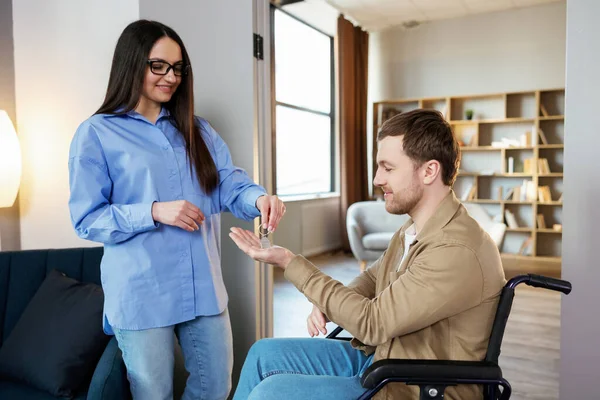 stock image Man in wheelchair extend her hand palm up to side. Woman giving key with keychain from new house for handicapped.