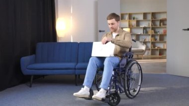 Tekerlekli sandalyedeki mutlu sakat adam dizüstü bilgisayar kullanıyor, evde çalışıyor. Genç engelli adamın işi var ya da internette okuyor.