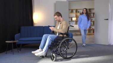 Kız arkadaşı dairede dolaşmasına yardım ederken tekerlekli sandalyedeki yakışıklı bir genç elinde cep telefonu tutuyor.