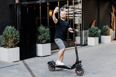 Elektrikli scooterlı yetişkin bir adam sokakta cep telefonuyla selfie çekiyor. Modern çevre dostu ulaşım kavramı