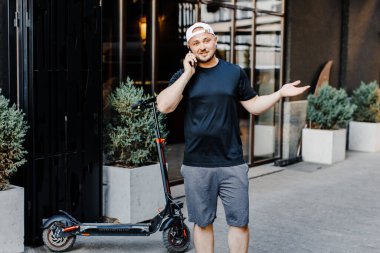 Caucasian young handsome man standing on street at electric scooter and talking on mobile phone.