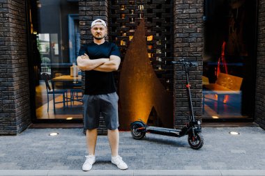 Sokakta elektrikli scooter 'ın yanında duran beyaz şapkalı ve günlük kıyafetleri olan yakışıklı bir genç. Ekolojik ulaşım kavramı