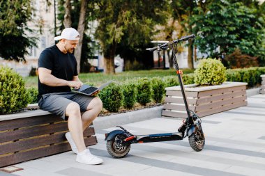 Şık bir adam, bir sokakta elektrikli scooterın yanında otururken dizüstü bilgisayarda çalışır. Sürdürülebilir ve çevre dostu yaşam tarzı kavramı