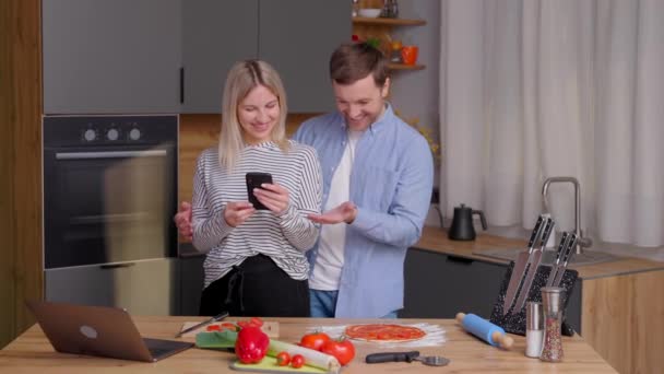 自宅のキッチンに立ち 携帯電話を使用している間 愛情のこもったカップルの料理サラダを一緒に笑顔 かわいいカップルの男と女は料理をしながら一緒に写真を撮る — ストック動画