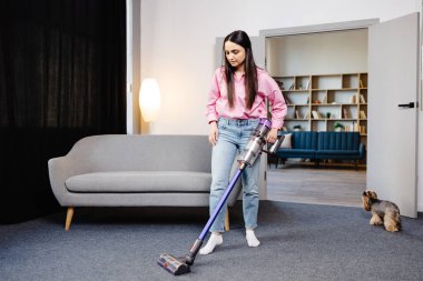 Genç bir kadın ev halısını temizlemek için kablosuz süpürge kullanıyor ve yakınlarda bir köpek koşuyor. Modern kolay temizlik.