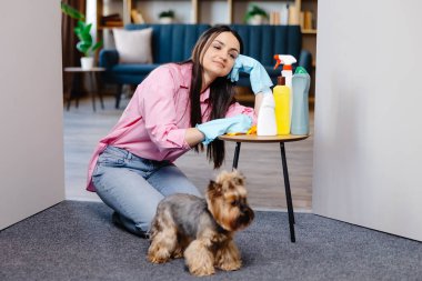 Evdeki temizlik malzemelerinin yanındaki masaya yaslanan genç bir kadın ve küçük köpeği onun yanında duruyor..