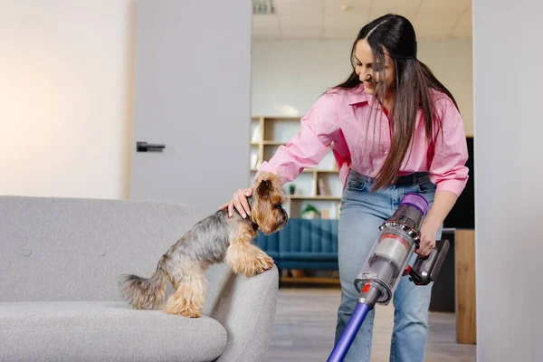 A cute young woman is vacuuming at home with a cordless vacuum cleaner and stroking her cute little dog with her hand