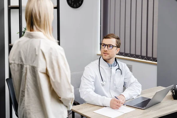 A young attractive doctor is having a consultation in his office. The doctor gives recommendations to his patient while sitting at the workplace.