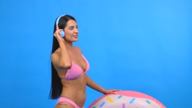 Pembe mayo giyen, kulaklıkla müzik dinleyen, elinde şişirilebilir halka tutan, dans eden, şarkı söyleyen ve mavi duvarda izole bir şekilde eğlenen mutlu genç bir kadın.