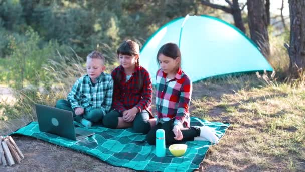孩子们在电脑上看表演 在森林里吃爆米花 男孩和女孩都在帐篷里休息 孩子们坐在草地上的格子花上 — 图库视频影像