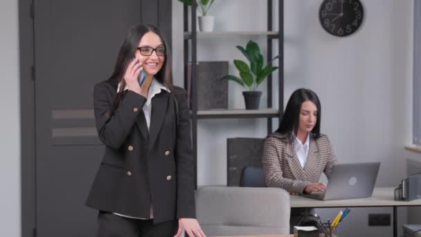 デスクに座っている間に携帯電話で話すジャケットを身に着けている魅力的な若いビジネス女性オフィスで同僚の近くのノートブックを使用して — ストック動画
