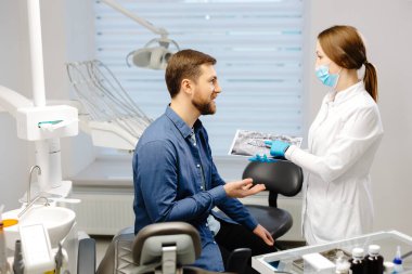 Young attractive man visiting dentist, sitting in dental chair at modern light clinic. Young woman dentist holding x ray image