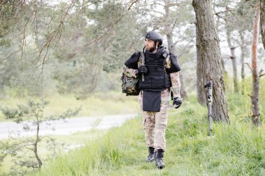 Üniformalı ve taktik yelekli genç asker ormanda çalışıyor ve geçici bir orman üssünde harekete geçmeye hazırlanıyor. Bir adam bölgeyi mayınlardan arındırmak için çalışır..