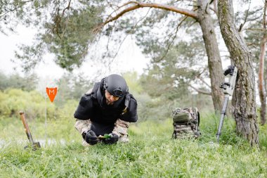Askeri üniformalı ve kurşun geçirmez yelek giyen bir adam ormanın mayınlarını temizlemek için çalışıyor. Bir adam kırmızı bir iz bırakarak tehlikeye karşı uyarır..