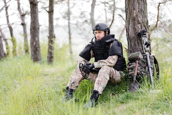 Un hombre con uniforme militar y chaleco antibalas trabaja en el bosque con  un detector de metales.
