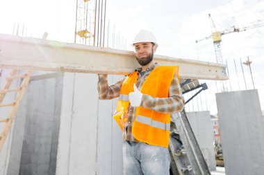 İnşaat sırasında inşaat alanında kereste taşıyan kasklı ve yelekli bir adam..