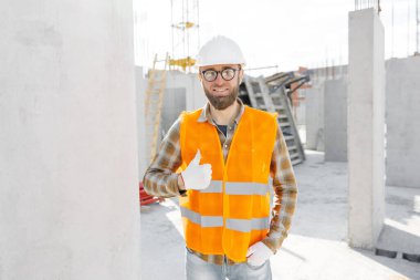 İnşaat işçisi, miğfer ve yelek giyen ustabaşı inşaat halindeki bir binada duruyor ve el hareketi gösteriyor..