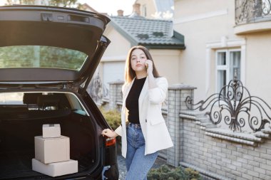 Young business woman standing at the car with parcels, coming home by car. The girl is standing and talking on mobile phone near the car.