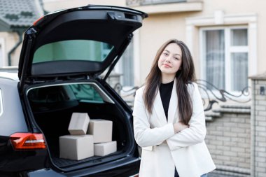 Genç bir kız arabanın bagajının yanında duruyor, kameraya bakıyor ve gülümsüyor..