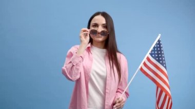 Gülümseyen mutlu genç kadın Amerikan bayrağı taşıyan günlük kıyafetler giyiyor ve mavi arka planda izole edilmiş kameraya bakıyor..