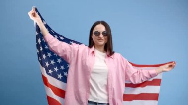 Gülümseyen mutlu genç kadın Amerikan bayrağı taşıyan günlük kıyafetler giyiyor ve mavi arka planda izole edilmiş kameraya bakıyor..