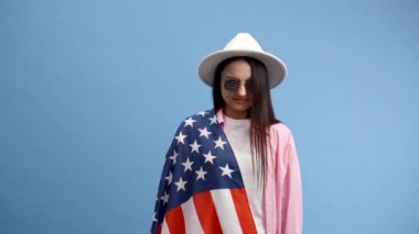 Gülümseyen mutlu genç kadın günlük kıyafetler giyiyor ve elinde Amerikan bayrağı olan beyaz şapka takıyor ve mavi arka planda izole edilmiş kameraya bakıyor..