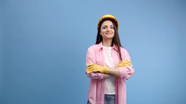 一个戴着黄色头盔和工作手套的年轻女子站在工作室的蓝色背景上 双手交叉在胸前 看着摄像机 — 图库视频影像