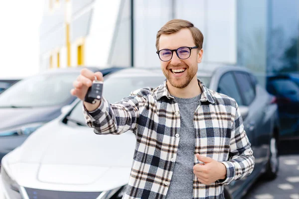 Emotional guy holding key in hand and smiling at camera. Man showing key from his new electric car. Young man buying auto at dealership salon.