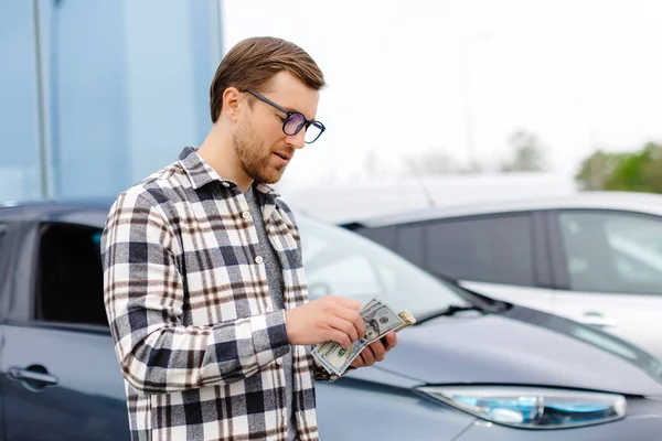Young man holding money in hand standing front car and prepare pay by installments - insurance, loan and buying car concept insurance. Buy and down payment a car.