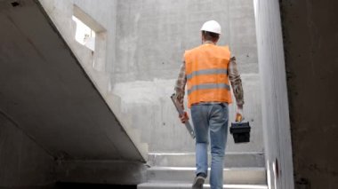 Tamirci, koruyucu miğfer ve yelek giyen ustabaşı inşaat alanında duruyor ve elinde cetvel ve alet çantası tutuyor.
