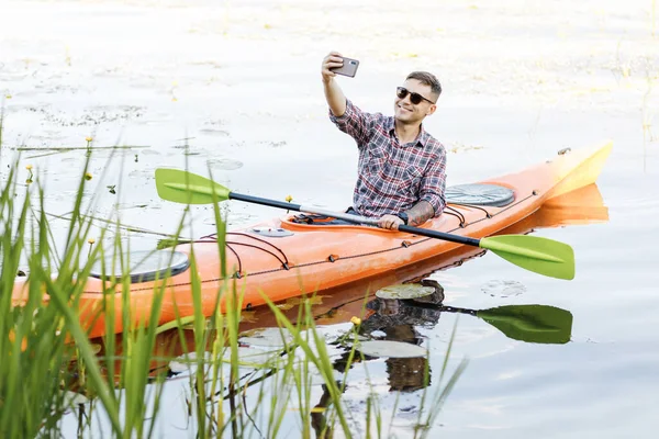 在河里划船 年轻的白人男子坐在皮划艇上 拿着手机自拍 水娱乐的概念 — 图库照片