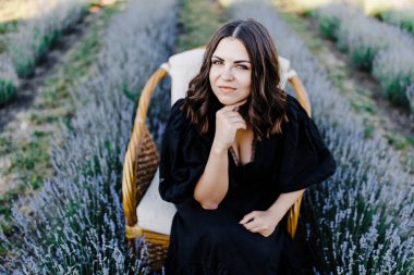 Attractive young woman in black dress and sitting in chair surrounded by lavender field.