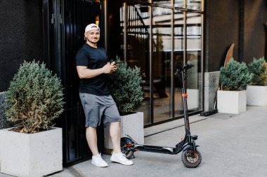 Modern binanın yanındaki caddede elektrikli scooterlı akıllı telefon kullanan genç adam.