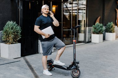 Elinde dizüstü bilgisayar olan yakışıklı serbest çalışan ve caddedeki elektrikli scooter 'da konuşan.