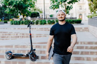 Elinde dizüstü bilgisayarı olan yakışıklı bir adam elektrikli scooter 'ın yanında duruyordu. Aktif yaşam tarzı, sırt çantalı gezgin. Ekoloji ve temiz ekolojik ulaşım biçimi