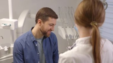 Young attractive man visiting dentist, sitting in dental chair at modern light clinic. Young woman dentist holding x ray image.