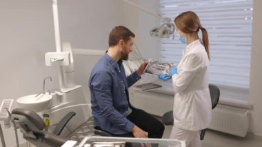 Young attractive man visiting dentist, sitting in dental chair at modern light clinic. Young woman dentist holding x ray image.