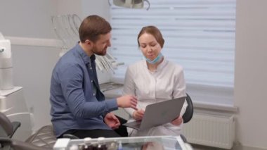 A handsome young man is talking to a female doctor at a dental appointment in a bright, beautiful office. The dentist explains to the patient and shows everything on the laptop