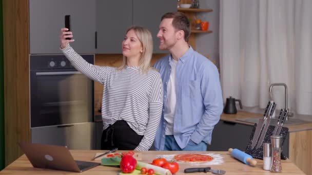自宅のキッチンに立ち 携帯電話を使用している間 愛情のこもったカップルの料理サラダを一緒に笑顔 かわいいカップルの男と女は料理をしながら一緒に写真を撮る — ストック動画