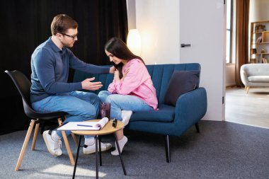 Kadın psikologla seans yapıyor, klinikte kanepede oturuyor. Kadın çok üzgün, psikoterapist ona yardım etmeye çalışıyor, moral desteği için omzunu tutuyor..