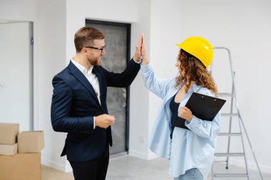 Miğferli kadın tasarımcı, elinde belgelerle odada dururken evden genç iş adamına anahtar veriyor..