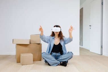 VR gözlüklü genç bir kadın modern teknolojiyi kullanarak oda tasarımı yapıyor, ellerini havada hareket ettiriyor, yenileme sırasında yeni bir dairede duruyor. Sanal gerçeklik ve onarım kavramı.