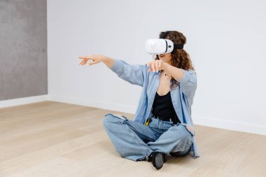 VR gözlüklü genç bir kadın modern teknolojiyi kullanarak oda tasarımı yapıyor, ellerini havada hareket ettiriyor, yenileme sırasında yeni bir dairede duruyor. Sanal gerçeklik ve onarım kavramı.
