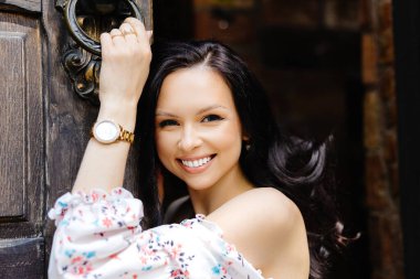 Genç ve güzel bir kadın açık eski bir kapının yanında duruyor, gülümsüyor ve kameraya bakıyor..