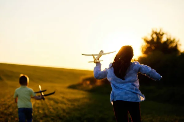 stock image Running boy and girl holding two yellow and blue airplanes toy in the field during summer sunset