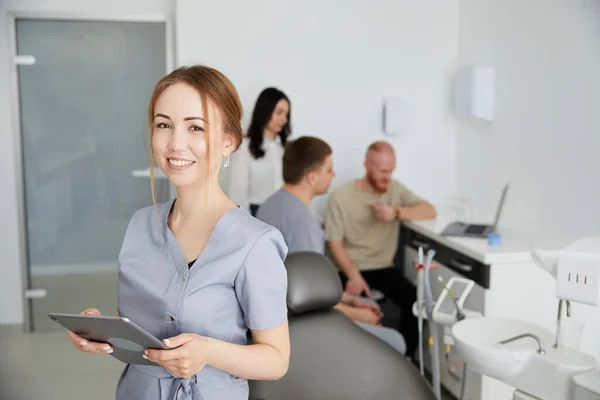一位年轻漂亮的女牙医站在办公室中央 一位同事正在为病人提供建议 为病人入院作准备 — 图库照片