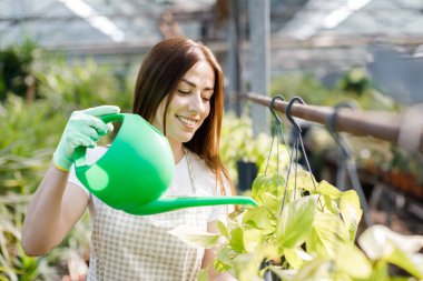Genç bir kadın büyük bir seranın ortasında duruyor ve bir su bidonundan saksıları döküyor..