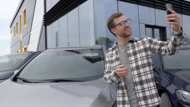 新車を買ってからスマホで自撮りしている幸せな男 携帯電話上のビデオリンク経由で購入した車を示す男 — ストック動画