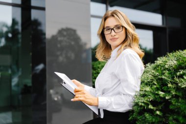 Kendine güveni tam genç bir kadın, resmi olarak giyinmiş, iş merkezinin arka planında oturuyor, elinde belgelerle bir klasör tutuyor, saate bakıyor..